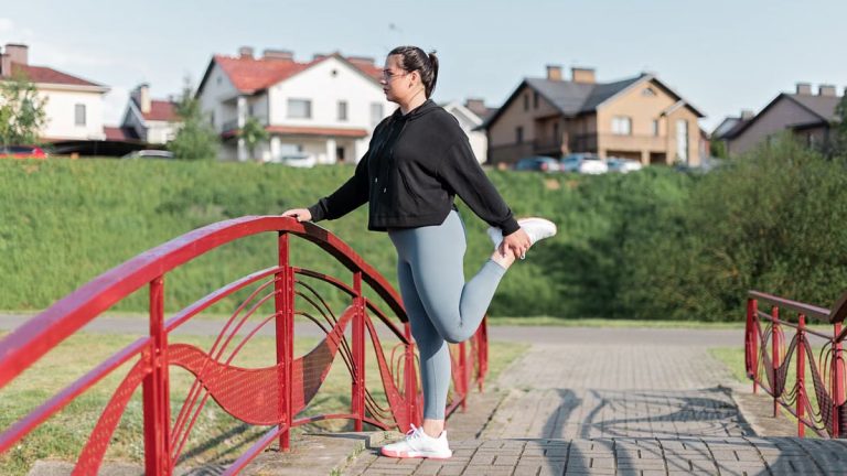 A woman in active wear performs a stretching exercise on a red bridge in a suburban park setting.